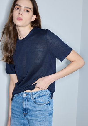 Jonge vrouw met lang bruin haar die een donkerblauw T-shirt met korte mouwen en lichtblauwe spijkerbroek draagt, en de zoom van het T-shirt bij de taille vasthoudt.