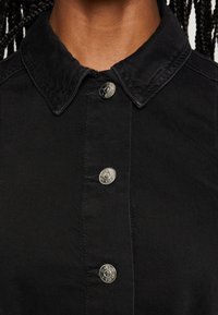 Lähikuva mustasta kauluspuserosta, jossa on metalliset painonapit, joiden päällä on teksti "ONLY", henkilöllä, jolla on tummat letitetyt hiukset.