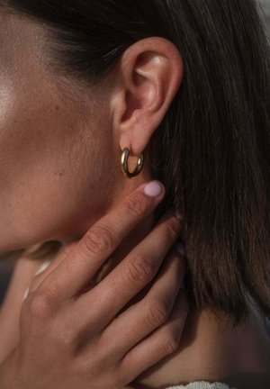 Nahaufnahme eines Ohrs mit kleinem goldenen Creolen-Ohrring und einer Hand, die sanft den Hals und die Kieferlinie berührt, braune Haare sichtbar.