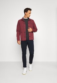 Röd bomulljacka med knäppning, två framfickor, lager över en mörkgrå tröja, i kombination med marinblå byxor och vita sneakers.