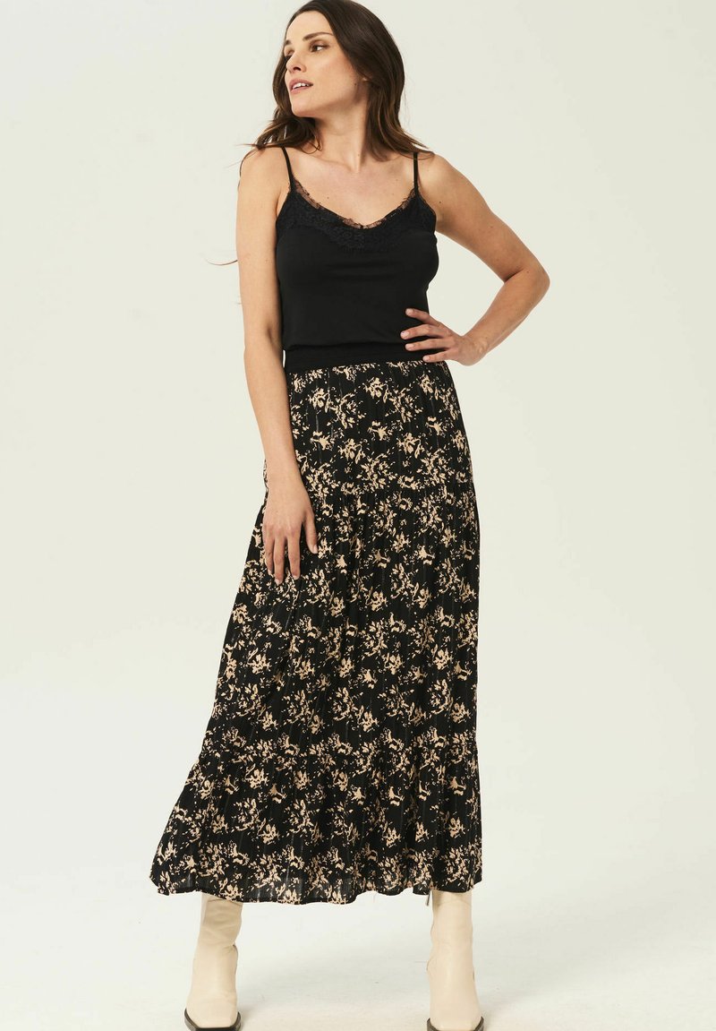Camisole noir à dentelle, assorti d'une longue jupe noire à motifs floraux avec des accents beiges et un design plissé, porté avec des bottes blanches.