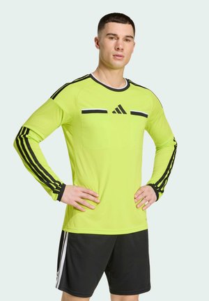 Hombre con camiseta deportiva de manga larga de color amarillo brillante con rayas negras y pantalones cortos negros, de pie con las manos en las caderas contra un fondo pálido.