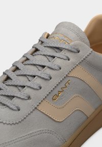 Αθλητικό παπούτσι από ανοιχτό γκρι σουέντ με μπεζ δερμάτινες λεπτομέρειες, γκρι κορδόνια και χρυσό λογότυπο "GANT" στο πλάι, με υφή στη σόλα από καουτσούκ.