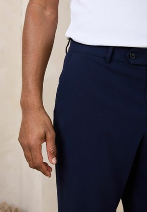 Linker Arm und Hand einer Person, die ein weißes ärmelloses Shirt und marineblaue Hose trägt, vor neutralem Hintergrund.