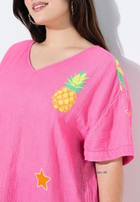 Różowa, oversize'owa koszulka z dekoltem w serek, z teksturowanej tkaniny, z żółtą grafiką ananasa i pomarańczowymi gwiazdkami na dolnej części przodu.