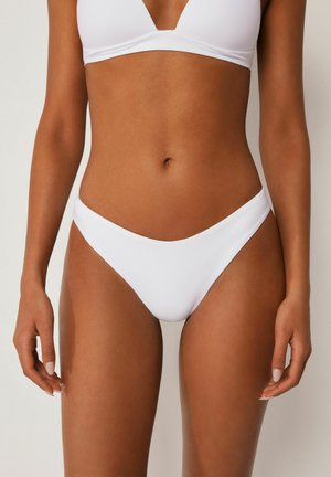 Frau trägt ein schlichtes weißes Bikini-Set, steht mit entspannten Armen seitlich an einem neutralen Hintergrund.