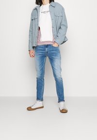 Giacca di jeans azzurro chiaro sopra un maglione bianco e rosa, abbinata a jeans azzurro chiaro e scarpe da ginnastica bianche con dettagli marroni.