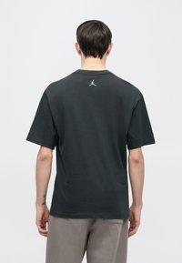 Juodas medvilninis trumpomis rankovėmis marškinėlis su mažu baltu Air Jordan logotipu viršutinėje nugaros dalyje. Lykus audinys ir laisvas siluetas.