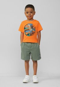 Oranje T-shirt met grafische print van palmbomen en een strandscène; gecombineerd met groene shorts; witte sneakers; kind staat met handen in de zakken.