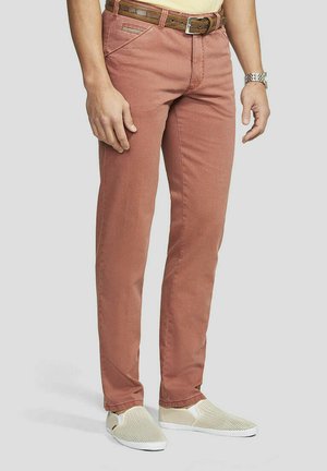 Man draagt roestkleurige broek, bruine riem, zilveren horloge en witte instappers, staand tegen een effen achtergrond.