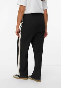 Pantalons noirs à coupe décontractée avec une jambe large, une taille élastique et une bande latérale beige contrastante. Portés avec des baskets noires.