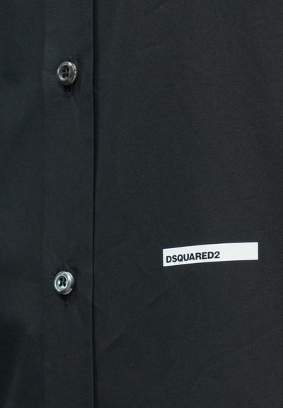 Sort skjorte med knapper, to synlige knapper og en hvid rektangulær label på højre side med teksten "DSQUARED2" i sorte bogstaver.