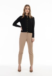 Cardigan nero a maglia con bottoni dorati, abbinato a pantaloni beige su misura. La modella indossa tacchi a stampa leopardata e ha i capelli lisci lasciati sciolti.