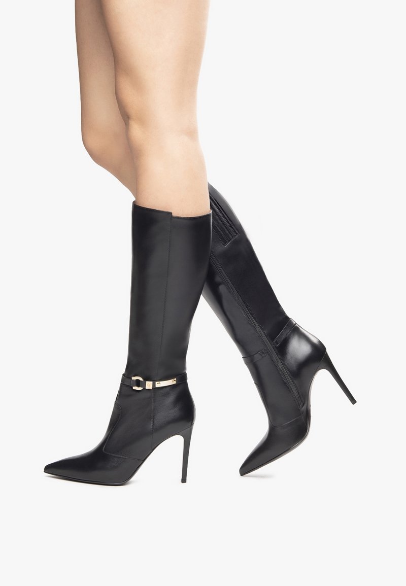 Jambes portant des bottes en cuir noires montantes jusqu'aux genoux, avec des bouts pointus, des talons aiguilles, et une boucle dorée décorative sur fond blanc.