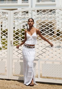 Robe maxi blanche en couches avec des fines bretelles et une texture froncée. Accentuée par une ceinture décorative avec des détails métalliques. Sandales noires aux pieds.