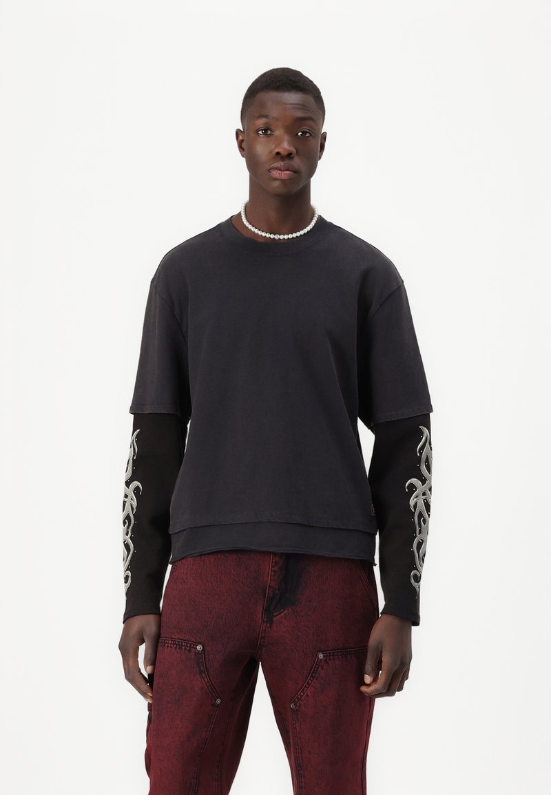 Black short-sleeve t-shirt with black long sleeves featuring silver embroidery. Paired with red textured pants, highlighting a modern design.
