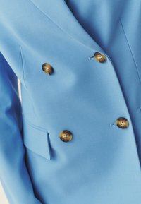Blazer bleu clair croisé à quatre boutons marron et deux poches à rabat, vu de la moitié supérieure du buste.