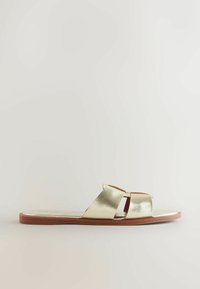 Sandalias planas metálicas doradas con dos correas cruzadas. Textura suave, punta abierta y suela plana de goma con un borde marrón contrastante.