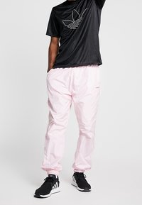 Chemise noire à manches courtes avec logo graphique blanc, associée à un pantalon de jogging rose clair, doté de poignets élastiques et de baskets noires.