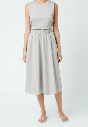 Femme portant une robe midi sans manches gris clair avec taille élastique et tongs blanches, debout devant un fond blanc uni.