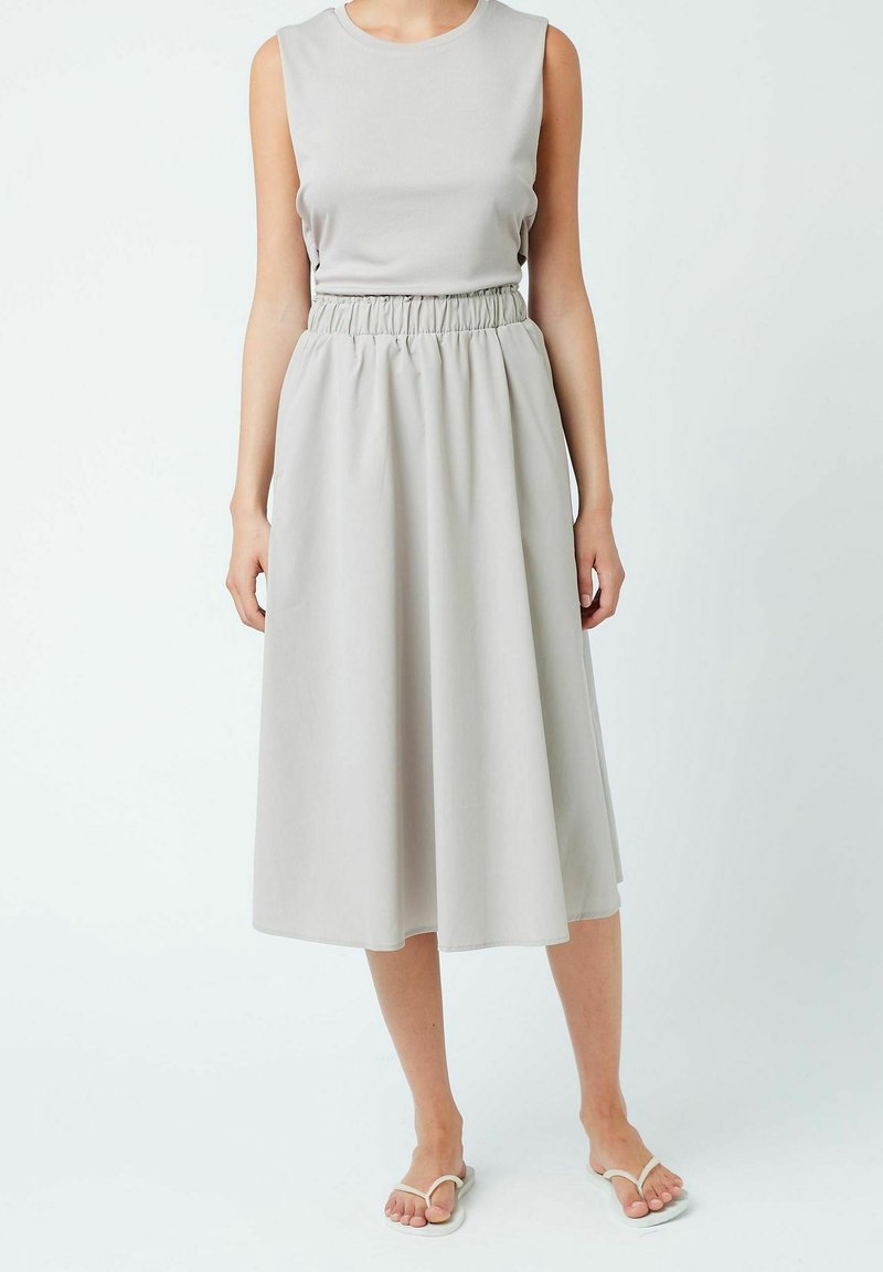 Femme portant une robe midi sans manches gris clair avec taille élastique et tongs blanches, debout devant un fond blanc uni.