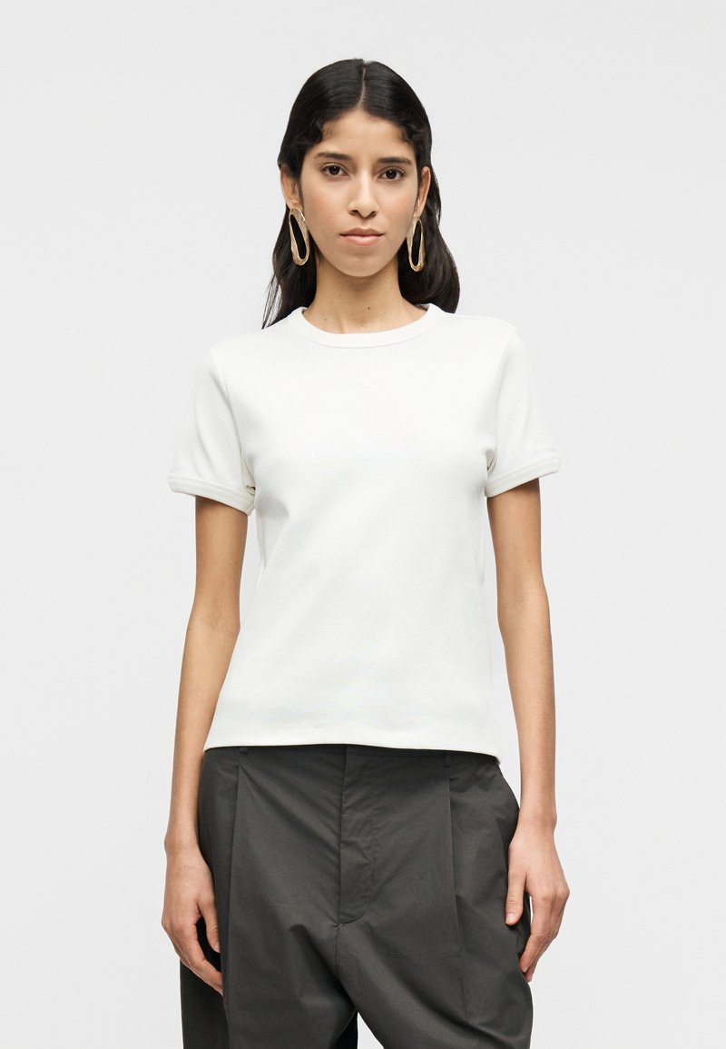 Λευκό κοντομάνικο μπλουζάκι με στρογγυλή λαιμόκ οψη και ρεβέρ στα μανίκια, κατασκευασμένο από λείο ύφασμα. Φορεμένο με ψηλόμεσο σκούρο παντελόνι.