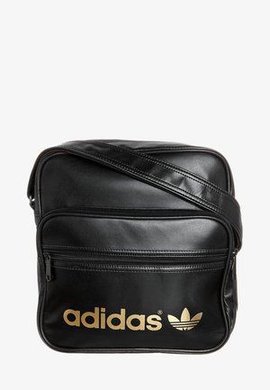 Sac bandoulière Adidas noir avec logo doré, poche zippée à l'avant et sangle d'épaule ajustable sur fond blanc.