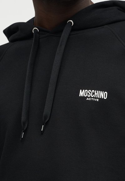 Svart hettegenser med tykke snorer og hvit "Moschino Active"-logo på venstre bryst, båret av en person med mørk hud.