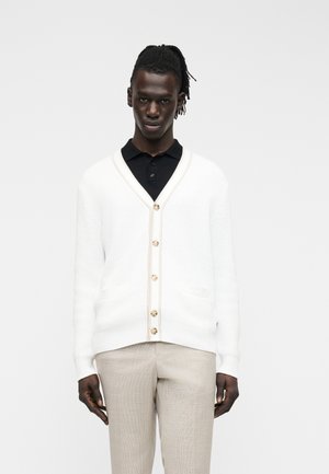 Man with dark skin and dreadlocks wears white buttoned cardigan over black shirt and beige pants, standing against a plain light background.