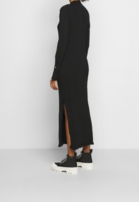 Vestito nero a coste con maniche lunghe, colletto alto, spacchi laterali e una finitura testurizzata. Abbinato a stivali neri chunky con suole bianche.