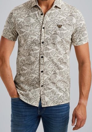 Uomo che indossa una camicia beige a maniche corte con motivo astratto grigio e piccolo logo sul petto, abbinata a jeans blu.