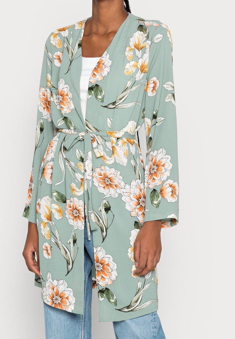 Kimono floral con fondo verde, que presenta flores naranjas y blancas. Tiene mangas largas y un cinturón de lazo en la cintura.