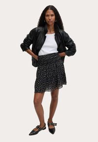 Frau mit langen lockigen Haaren, die eine schwarze Lederjacke, ein weißes Oberteil, einen schwarzen Blumenrock und schwarze spitze Ballerinas mit Schnallenriemen trägt, steht.