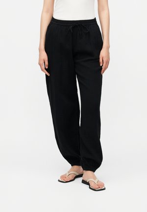 Mujer con pantalones negros sueltos con cordón y puños elásticos, y sandalias beige de uña, de pie frente a un fondo blanco liso.