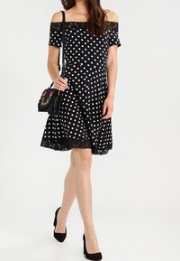 Svart av-axlar klänning med vita prickar, spetsdetalj vid fållen, utsvängd kjol, tillsammans med en svart axelremsväska och svarta klackar.