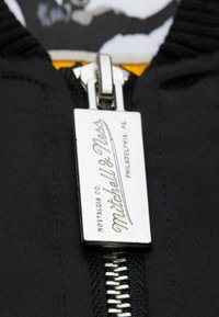 Schwarzes Kleidungsstück mit Reißverschluss und metallic Reißverschlusszug mit "Mitchell & Ness" Logo. Glatte Stofftextur mit detaillierten Nähten.
