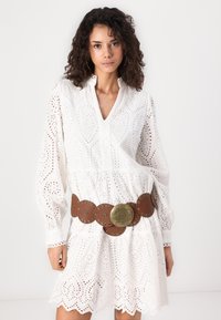 Vit broderie anglaise klänning med långa ärmar, V-ringning och vågformad nederkant, parat med ett brett bälte i brunt läder med cirkulära detaljer.