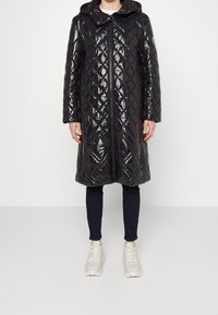 Cappotto trapuntato nero e lucido con cappuccio, caratterizzato da un motivo a rombi diagonale, chiusura con zip e tasche laterali, indossato sopra pantaloni scuri e sneakers.