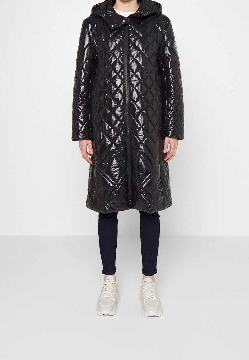 Cappotto trapuntato nero e lucido con cappuccio, caratterizzato da un motivo a rombi diagonale, chiusura con zip e tasche laterali, indossato sopra pantaloni scuri e sneakers.