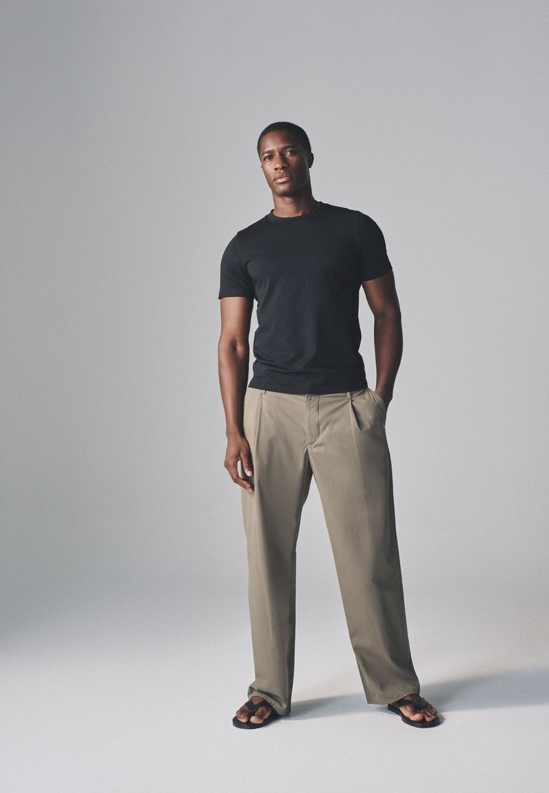 Homme debout, une main dans la poche, portant un t-shirt noir, un pantalon beige et des sandales noires sur un fond gris uni.