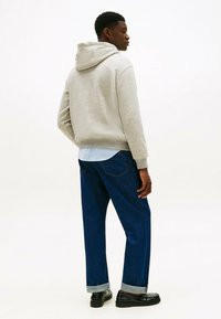 Uomo che indossa un hoodie grigio chiaro, jeans blu risvoltati, scarpe nere, in piedi e che guarda a destra su uno sfondo bianco semplice.