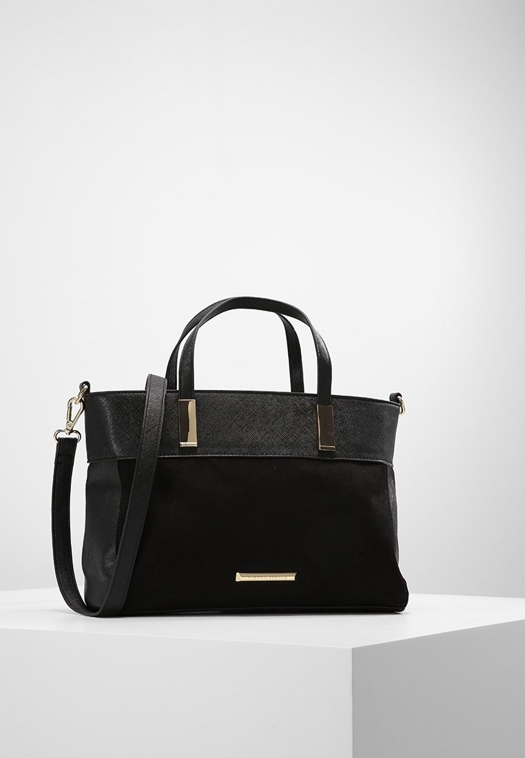 Sac à main noir avec deux poignées courtes, une bandoulière réglable et des accents métalliques rectangulaires dorés, posé sur une surface blanche.