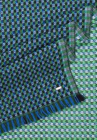 Tissu tissé à motifs bleus et verts avec des bords frangés, plié sur un textile tissé similaire en vert et bleu.