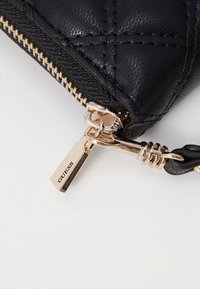 Portafoglio in pelle nera impunturata con zip e accessori in tonalità oro. Presenta un'etichetta incisa con "GUESS" e una finitura testurizzata.