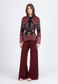 Giacca nera con intricata ricamo floreale in rosso e oro, abbinata a pantaloni bordeaux a gamba larga. La giacca presenta maniche lunghe e una silhouette aderente.
