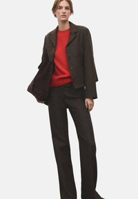 Blazer marron avec deux poches avant, associé à un pull tricoté rouge vif et un pantalon marron assorti, tenant un sac marron foncé.