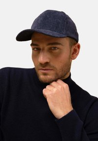 Námořnická kroucená čepice s zakřiveným kšiltem. Žebrovaná textura a strukturovaný design. Nosená nad černým pleteným rolákem, který zvýrazňuje rysy čepice.