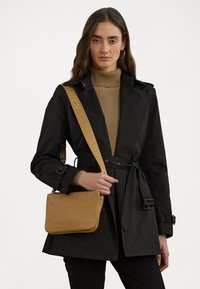 Manteau noir en trench avec une taille ceinturée, col roulé côtelé beige, et un sac bandoulière beige avec fermeture éclair et logo en relief.