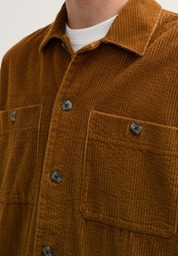 Brown corduroy shirt featuring a textured finish, two chest pockets, and black buttons for closure. Close-up view of collar and fabric.