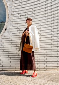 Frau in braunem Kleid und weißem Blazer steht vor einer Backsteinmauer, hält eine beigefarbene Handtasche und trägt rote Plateau-Sandalen.